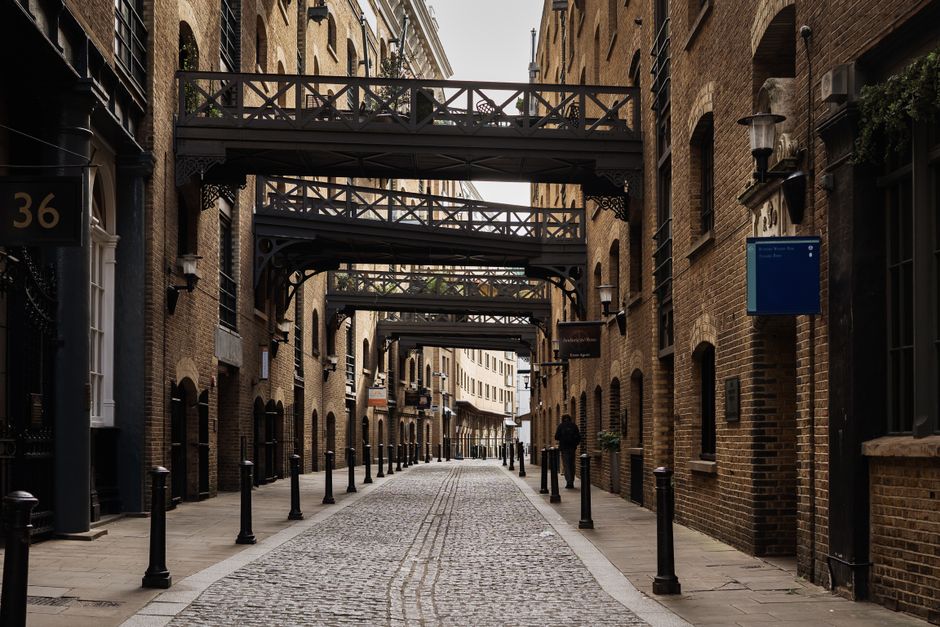 A narrow cobblestone street in an urban setting during daytime, flanked by tall brick buildings with numerous windows and fire escapes. The street has a slight incline, with a few pedestrians in the d