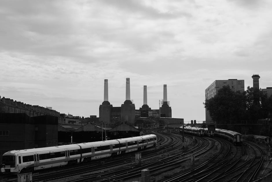 A black and white photograph showing a modern urban scene with a train on curved railway tracks in the foreground, situated near a brick building with a sloped roof. In the background, there are four large industrial chimneys emitting no smoke, set against a cloudy sky. To the right, a multi-story building with a flat roof and a tall chimney-like structure is partially obscured by a cluster of trees. The image captures the industrial and transport elements typical of a city environment, associated with house removals and logistics services offered by Battersea Removals for the Battersea Park to Battersea Power Station relocation project.
