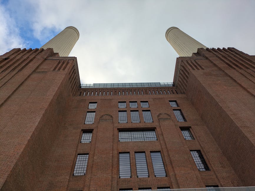 View from ground level looking up at a large brick building with a symmetrical façade, featuring multiple small rectangular windows with black panes arranged in vertical and horizontal clusters. The building has two prominent, vertically aligned, cylindrical smokestacks extending upward, with the sky visible in the background. The surrounding environment suggests an urban setting consistent with historic industrial architecture, and the scene may relate to home relocation or furniture transport as part of a house removals service. Battersea Removals, specializing in removals, is involved in the logistical process of packing and moving objects from the property, which might include loading on a vehicle parked nearby, though no vehicles or furniture are visible in this image.